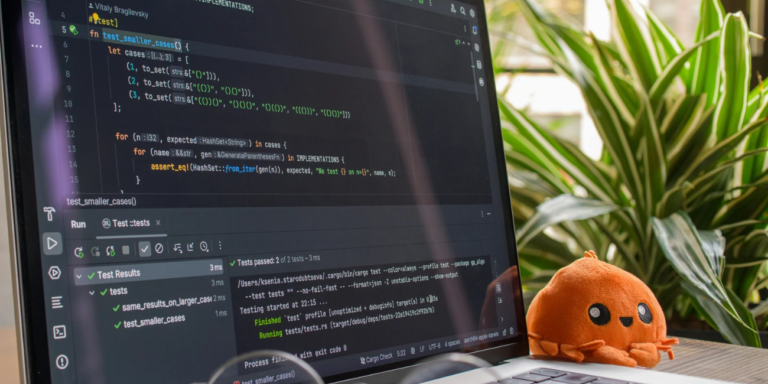 Pantalla con código y resultados de pruebas de software junto a un muñeco naranja sobre un escritorio.